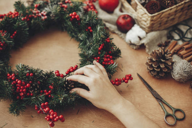 istock Hands making rustic christmas wreath with fir branches, red berries,pine cones,rope, scissors, cinnamon, cotton on rustic wooden background. Atmospheric moody image at holiday workshop 1075045230