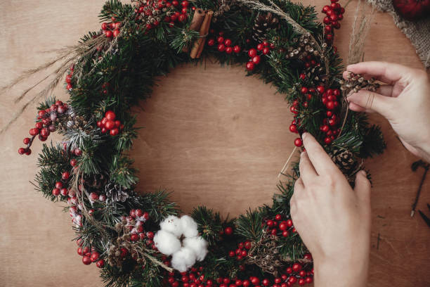 istock Hands making rustic christmas wreath, holding herbs at fir branches, red berries , pine cones, cotton on rustic wooden background. Atmospheric moody image at winter holiday workshop 1075045302