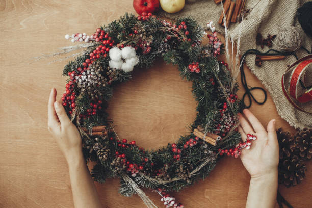 istock Rustic Christmas wreath. Hands holding fir branches, red berries and pine cones,rope, scissors, cinnamon, cotton on rustic wooden background. Atmospheric moody image at holiday workshop 1075046426