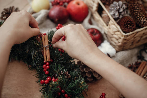 istock Hands making rustic christmas wreath, holding cinnamon at fir branches, red berries,pine cones,rope, scissors, cotton on rustic wooden background. Atmospheric moody image at holiday workshop 1075046750