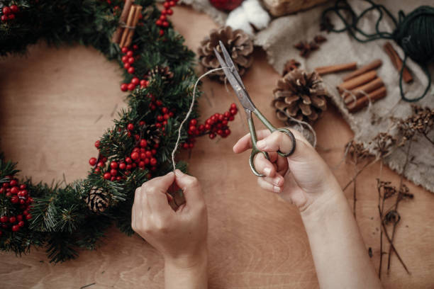 istock Hands making rustic christmas wreath, cutting rope with  scissors at fir branches, red berries,pine cones on rustic wooden background. Atmospheric moody image at holiday workshop 1075046920