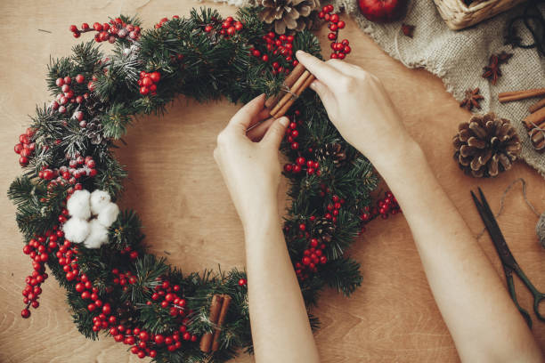 istock Hands making rustic christmas wreath, holding cinnamon at fir branches, red berries,pine cones,rope, scissors, cotton on rustic wooden background. Atmospheric moody image at holiday workshop 1075047006