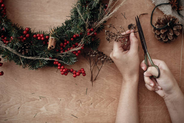 istock Hands making rustic christmas wreath, holding scissors at herbs, fir branches, red berries, pine cones on rustic wooden background. Atmospheric moody image at winter holiday workshop 1075047028