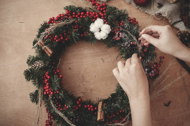 istock Hands making rustic christmas wreath, holding herbs at fir branches, red berries , pine cones, cotton on rustic wooden background. Atmospheric moody image at winter holiday workshop 1075047054