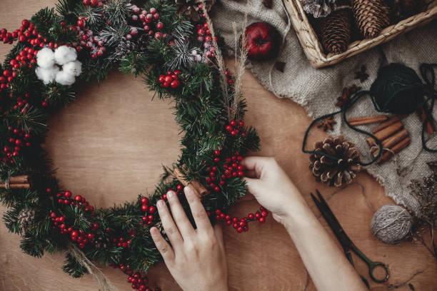 istock Rustic Christmas wreath flat lay. Hands holding fir branches, red berries and pine cones,thread, scissors, cinnamon, cotton on rustic wooden background. Making  wreath at workshop 1075047776