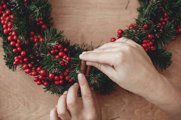 istock Hands making rustic christmas wreath, holding cinnamon at fir branches, red berries , pine cones, cotton on rustic wooden background. Atmospheric moody image at winter holiday workshop 1075048528