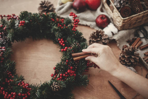 istock Hands making rustic christmas wreath, holding cinnamon at fir branches, red berries,pine cones,rope, scissors, cotton on rustic wooden background. Atmospheric moody image at holiday workshop 1075048682