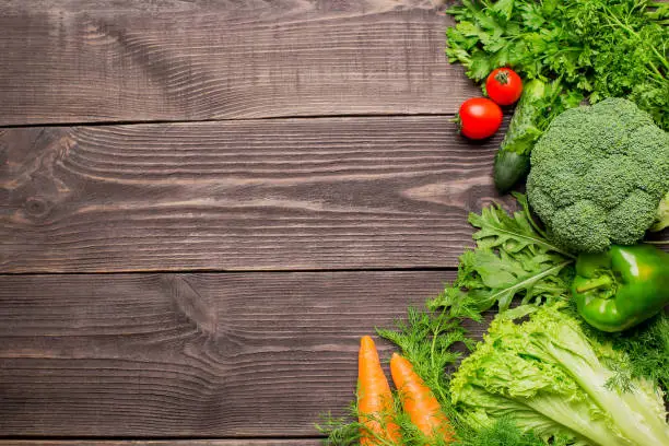 Frame of bright green and red fresh vegetables on wooden background, copy space. Pepper, broccoli, lettuce, arugula, parsley, carrot