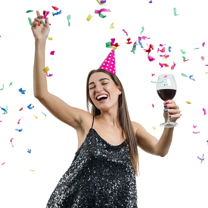 Graceful, smiling young brunette girl in party hat with glass of red wine dancing under confetti and laughing, white isolated background. Party, drinks, holidays and celebration concept