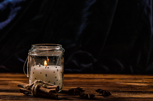 Close up. A white candle in a transparent glass jar is burning, cinnamon, anise are laid out around it. Rustic brown wooden table, dark blue background. Copy space.