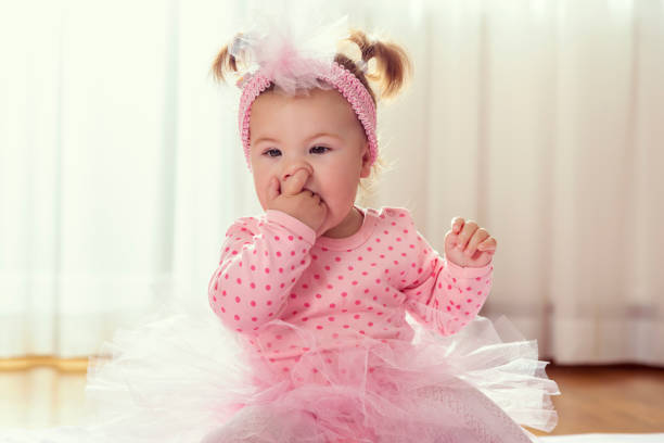Beautiful little baby girl sitting on the nursery floor, wearing pink tutu skirt, chewing her hands and making faces