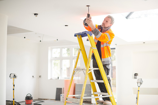 Full Length Portrait Of Electrician On Stepladder Installs Lighting To The Ceiling In Office