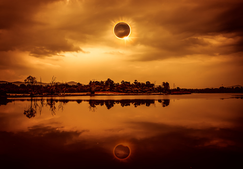 Amazing scientific natural phenomenon. The Moon covering the Sun. Total solar eclipse with diamond ring effect glowing on sky above silhouettes of trees, river area. Serenity nature background.