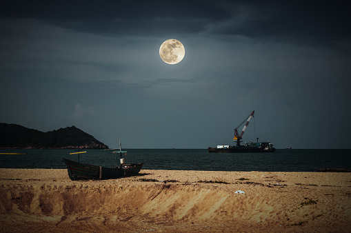 Beautiful night sky with bright full moon above cargo ship anchored in the sea. Traditional fishing boat parked on tropical beach. Outdoor at nighttime. The moon taken with my camera.