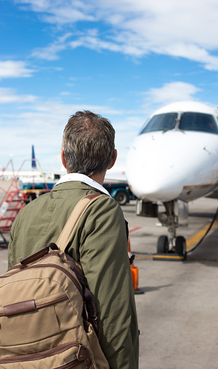 An active senior man stands with a backpack on the tarmac looking at a small plane. Shot at the Santa Fe, NM airport.