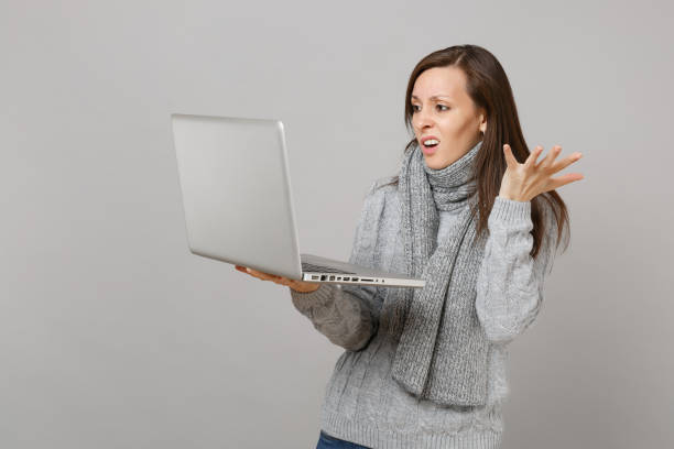 istock Perplexed young woman in gray sweater and scarf spreading hands, working on laptop pc computer isolated on grey wall background. Healthy lifestyle, online treatment consulting, cold season concept. 1075335738