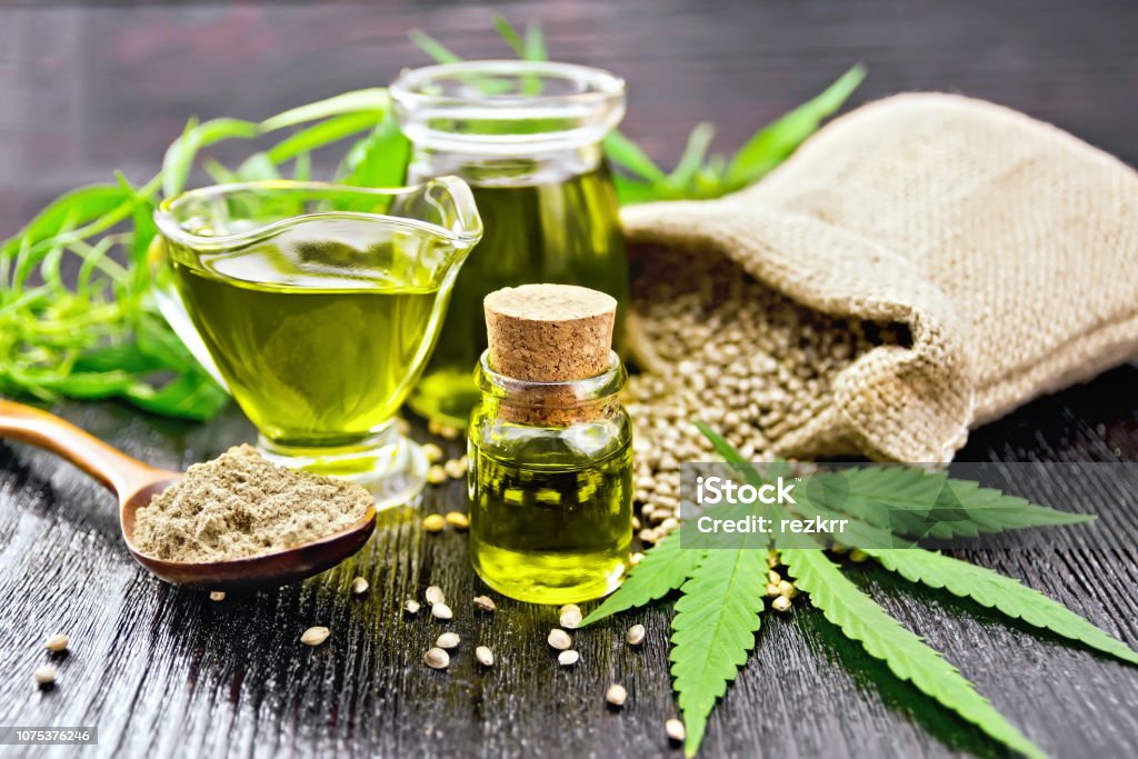 Oil hemp in two jars and sauceboat on wooden board Hemp oil in two glass jars and sauceboat with grain in the bag, leaves and stalks of cannabis, a spoon with flour on the background of wooden boards Cannabis Plant Stock Photo