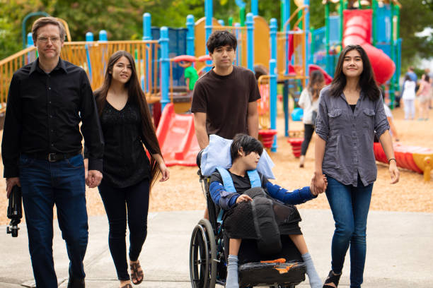 Famiglia che cammina davanti al parco giochi con figlio disabile in sedia a rotelle - foto stock