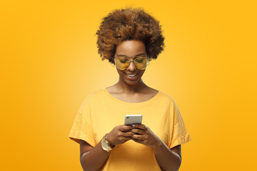 Young african american woman in yellow t-shirt and trendy eyeglasses looking at screen of phone and smiling nicely while chatting