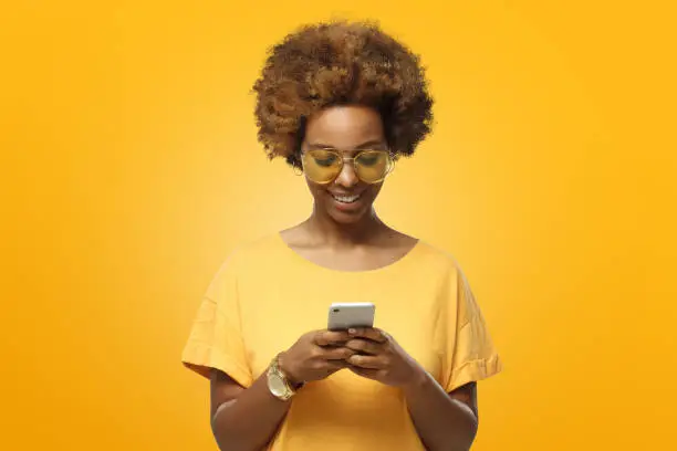 Photo of Young african american woman in yellow t-shirt and trendy eyeglasses looking at screen of phone and smiling nicely while chatting