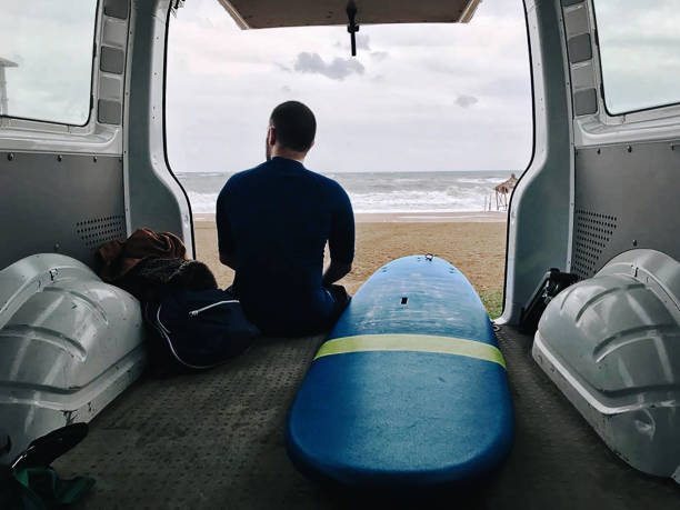 Young man behind van with his surfboard looking through beach and waves Young man sitting behind van with his surfboard, waiting for the waves an looking wavy sea. Cloudy and rainy gray weather outside. Getting away from it all. neoprene photos stock pictures, royalty-free photos & images