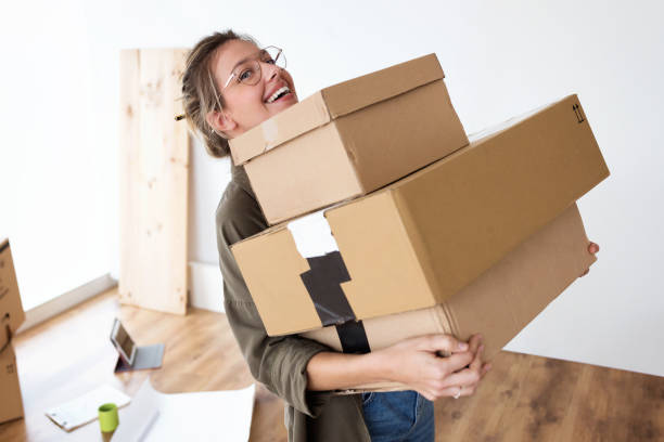 Pretty young woman carrying cardboard boxes into her new house while smiling at camera.