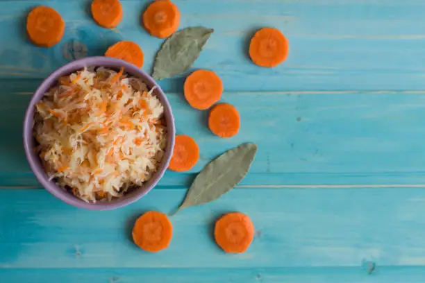 Photo of Photo of sauerkraut in a plate and round slices of carrots and bay leaf on a bright blue background.