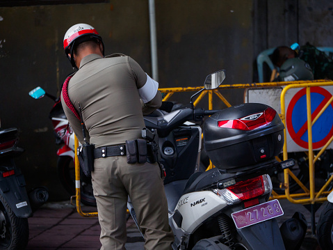 BANGKOK, THAILAND - JULY 30, 2018: A uniformed police officer issues a parking ticket to motorcycles parked in restricted areas near a barricade. Crime and law enforcement.