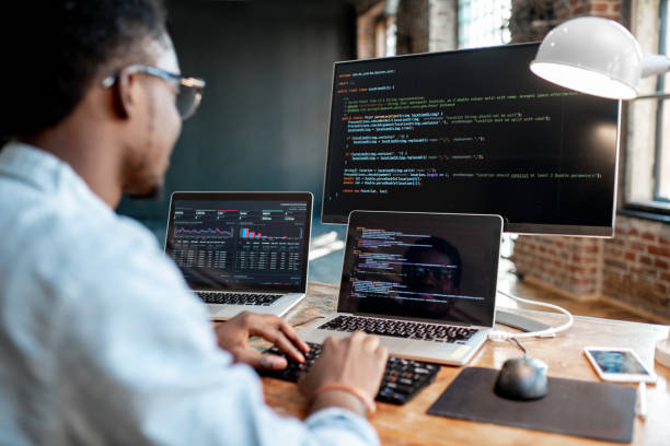 Programmer working with program code Young african male programmer writing program code sitting at the workplace with three monitors in the office. Image focused on the screen development stock pictures, royalty-free photos & images