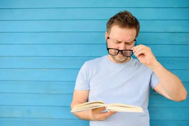 Portrait of mature man with big black eye glasses trying to read book but having difficulties seeing text because of vision problems. Problems disorder vision.