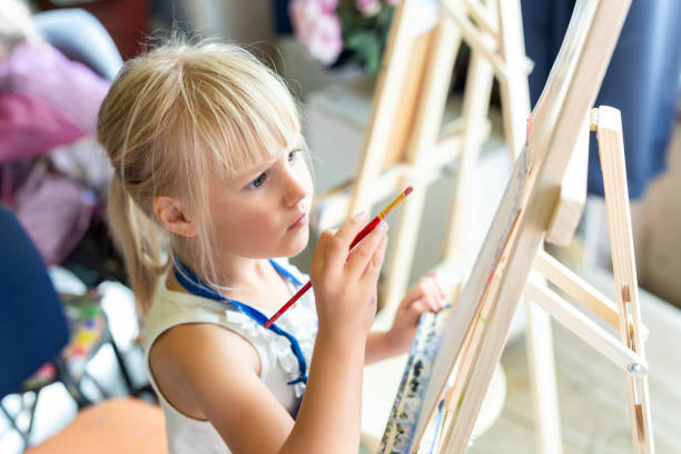 かわいいブロンドの笑顔の女の子はアート スタジオでワーク ショップ レッスンのイーゼルの絵画します。塗料で描いていて、手の楽しい子供持株ブラシ。子の開発コンセプト - artists canvas indoors childhood small ストックフォトと画像