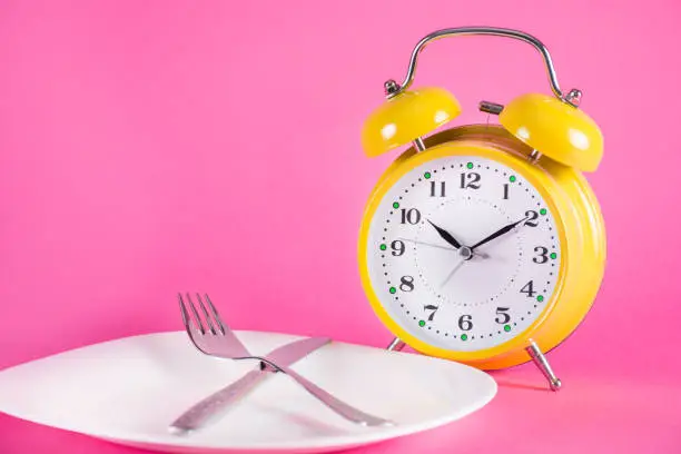 Old alarm clock and empty plate with fork and knife isolated on pink background. Food and Chrono nutrition diet concept. Close up, selective focus