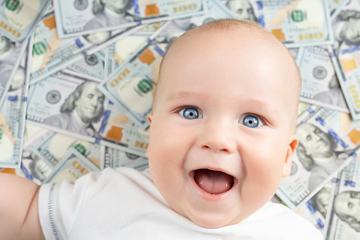 Cute happy baby boy smiling with hundred dollar bills background. Adorable kid having fun lying over american cash currency banknotes. Easy money and quick success business concept.
