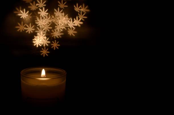 istock White aromatic glass candle with flame and snowflake shape bokeh on dark background for Christmas holiday. 1075777676