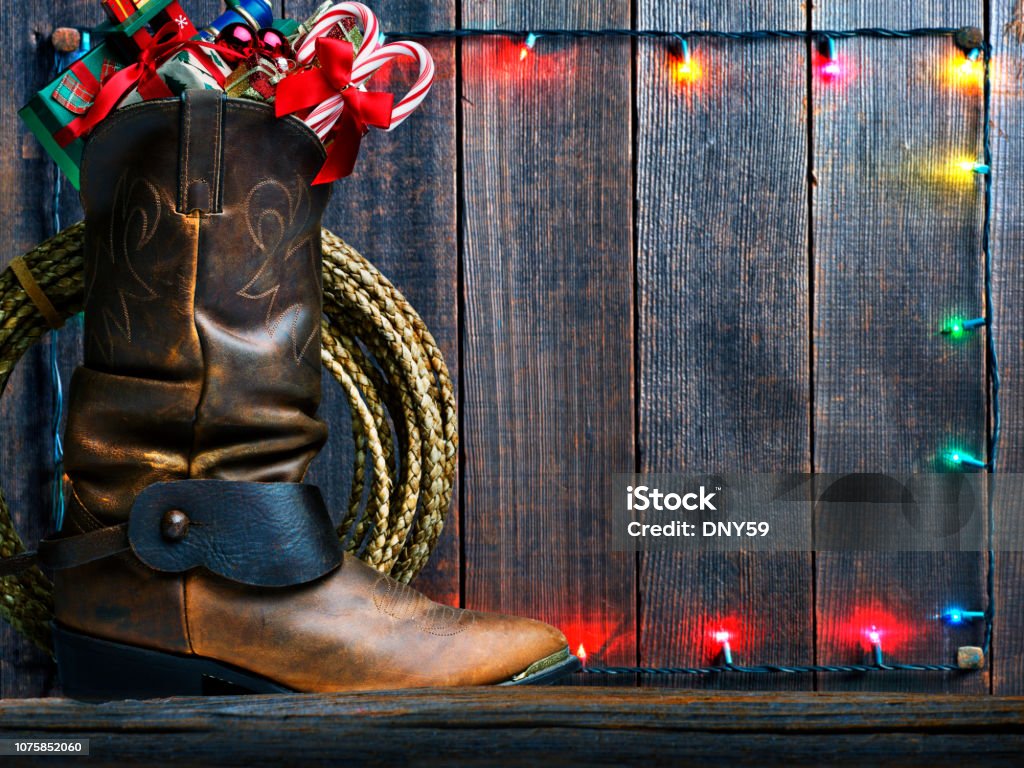 Cowboy Boot Stuffed With Christmas Presents An old cowboy boot is stuffed with small Christmas presents as it sits in front of  an old fence adorned with a strand of Christmas lights. Wild West Stock Photo
