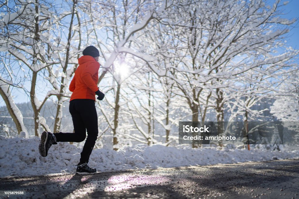 side view woman jogging running on road cold sunny winter day fit sporty female athlete 45 years old woman running jogging on road with slush on cold sunny sunshine winter day snow snow covered trees endurance training side view Winter Stock Photo