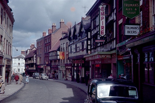 Shrewsbury, West Midlands, England, UK, 1960. Street scene at Old Potts Way in the medieval town of Shrewsbury. Furthermore: cars, half-timbered houses, locals, cinema, restaurant and advertising signs..