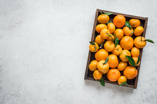 Tangerines (oranges, clementines, citrus fruits) with green leaves in box on white stone background, copy space.