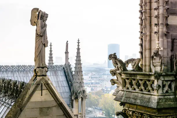 Stone statue of an angel with trumpet on the rooftop of Notre-Dame de Paris cathedral facing chimeras on the towers gallery with the buildings of the city below, vanishing in the mist.