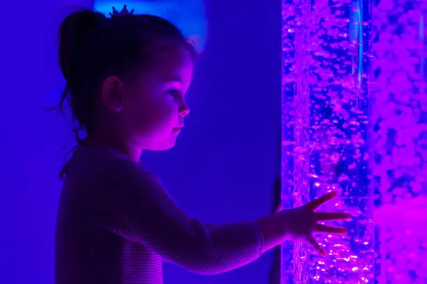 Photo of Child in therapy sensory stimulating room, snoezelen. Child interacting with colored lights bubble tube lamp during therapy session.