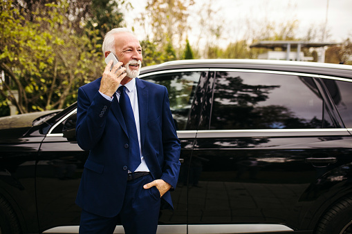 Senior adult driver standing and talking on the phone next to luxury car