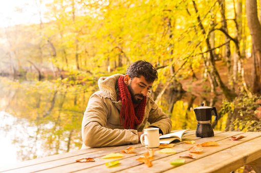 Young man reading book and drinking coffee while camping in the autumn nature