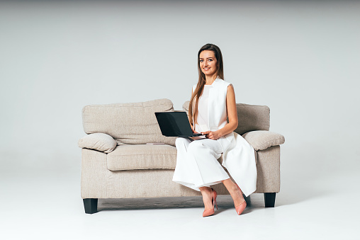 Modern businesswoman wearing white suit sitting on sofa and working on laptop over gray background. Beautiful female on a couch with a black laptop.