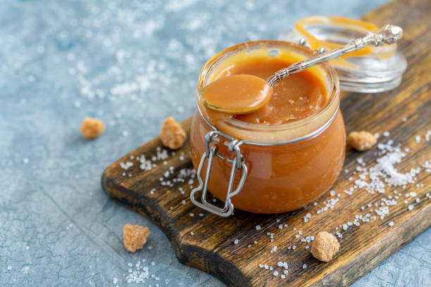 Glass jar with homemade salted caramel. Caramel sauce with sea salt in a small jar, brown cane sugar and a spoon with salt on an old wooden serving board, selective focus. caramel photos stock pictures, royalty-free photos & images