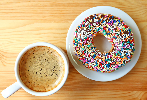 Top view of a cup of hot coffee and a donut topped with colorful sprinkles served on wooden table