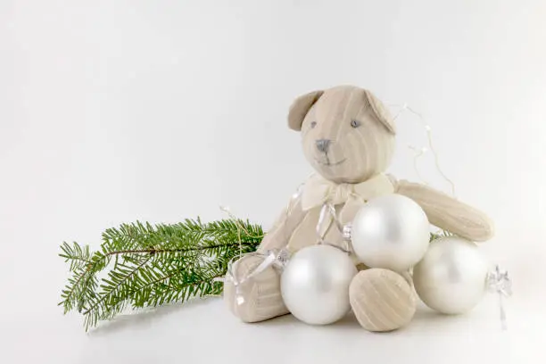 Teddy bear sitting with Christmas tree ornaments, on white background with green branches and fairy lights
