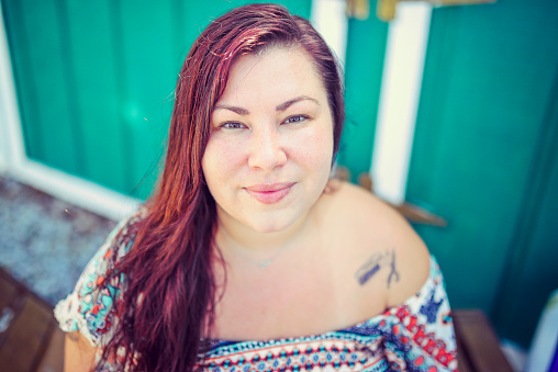 Portrait of a woman in her 30's with long dark hair and brown eyes. She wears an off shoulder top and has tattoos. She smiles (or smirks?) at the camera in a headshot. She is a mixed race Native American with Cherokee ancestry