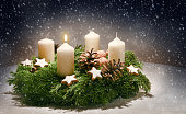 Advent wreath from evergreen branches with white candles, the first is burning for the time before Christmas, dark snowy background with copy space