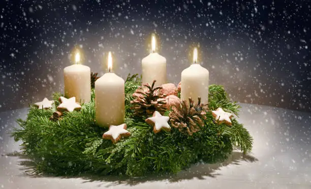 Advent wreath from evergreen branches with white candles, the fourth is burning for the time before Christmas, dark snowy background with copy space, selected focus