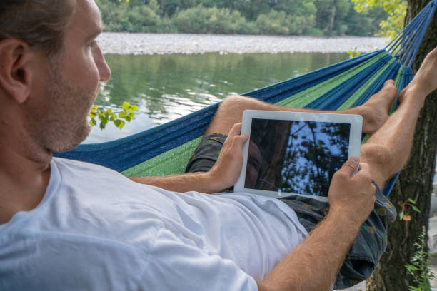 istock Young man using digital tablet on hammock, lying between trees by the river un Summer- People travel vacations technology on the go concept 1076372924